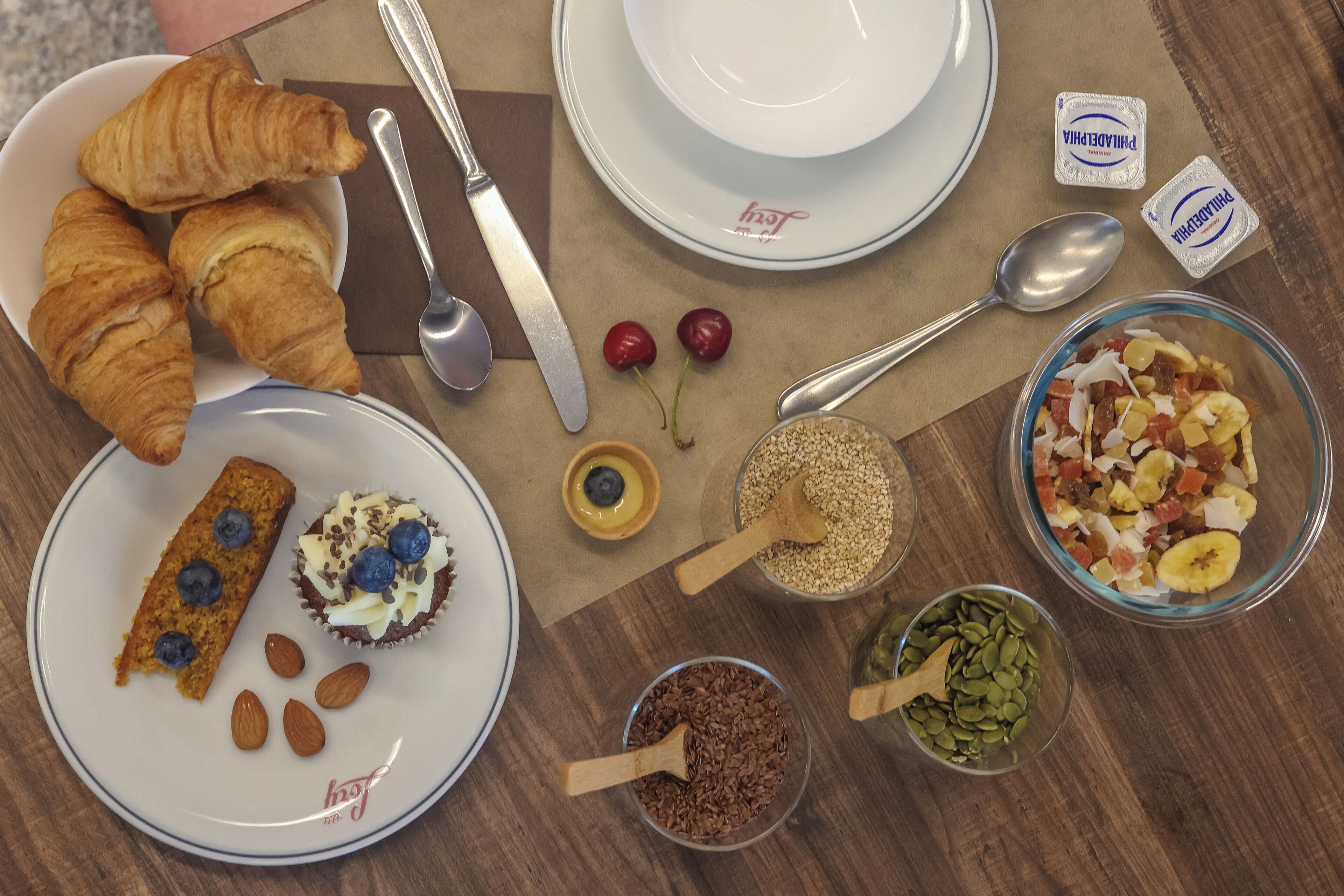A breakfast setting with croissants, a blueberry muffin, almonds, and various breakfast items on a wooden table.