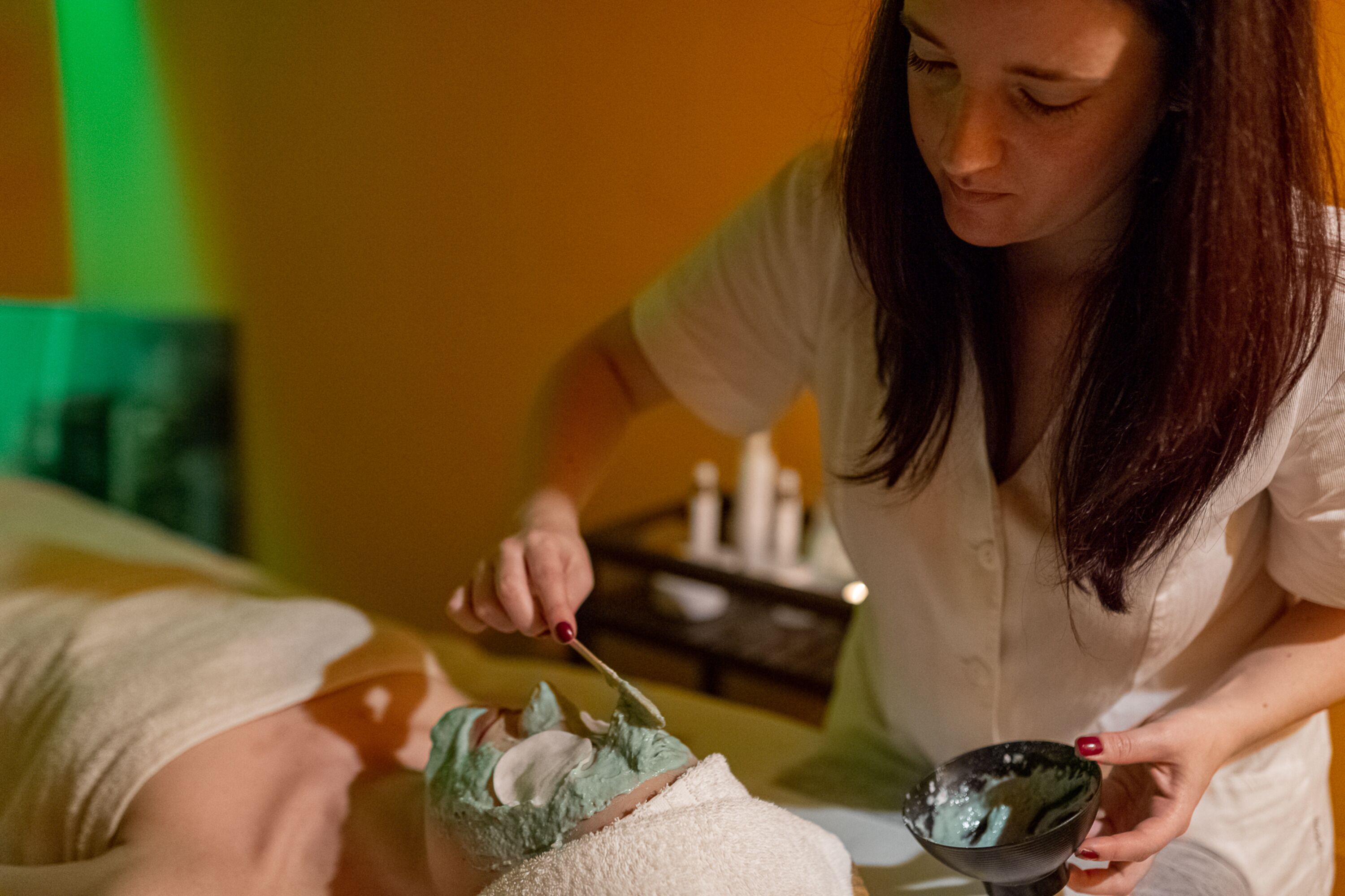 A person receiving a facial treatment in a room with a bed and a small table with bottles.
