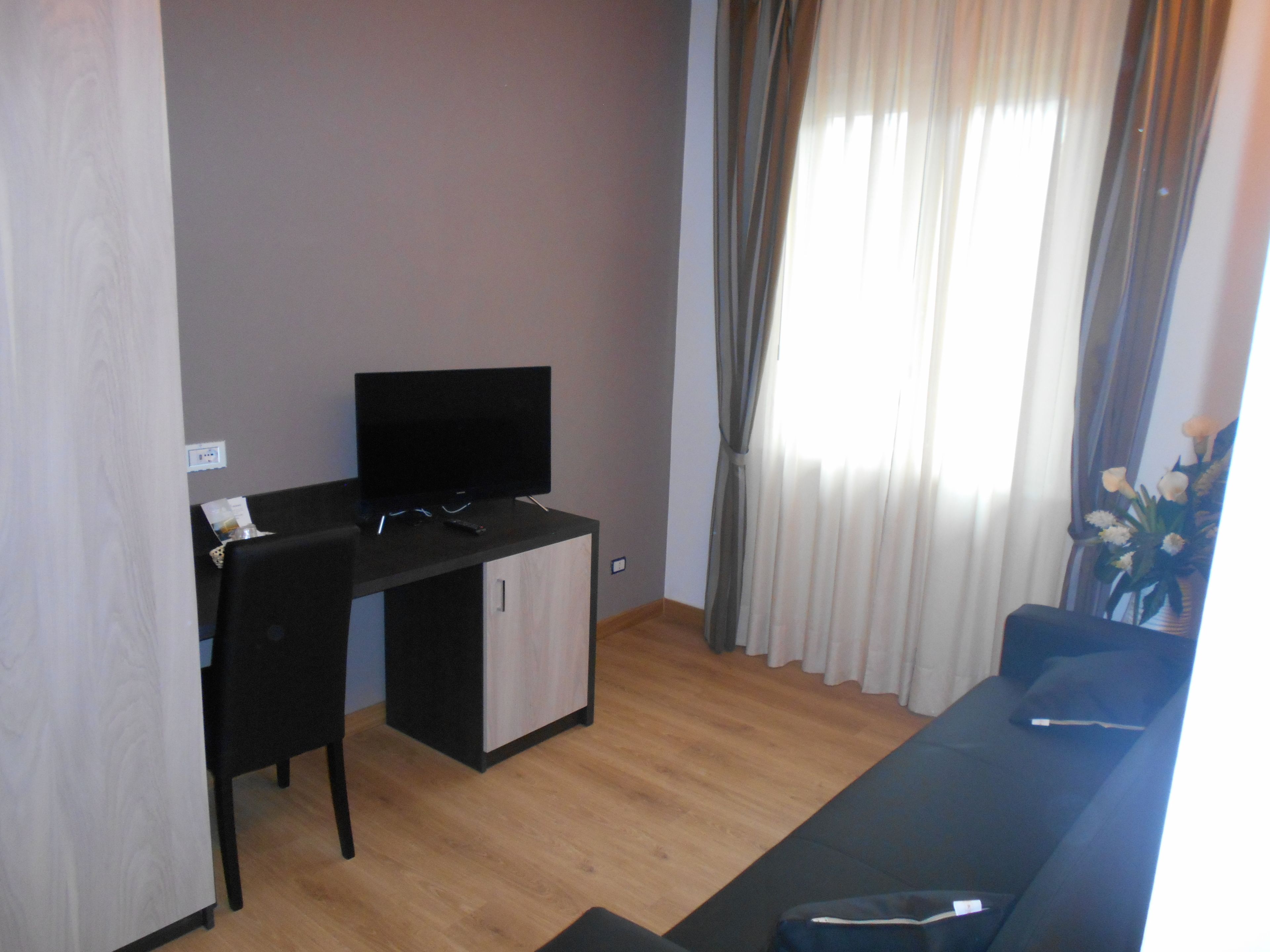 Un soggiorno moderno con una TV a schermo piatto su una scrivania nera, un pavimento in legno, un divano blu e una finestra con tende.