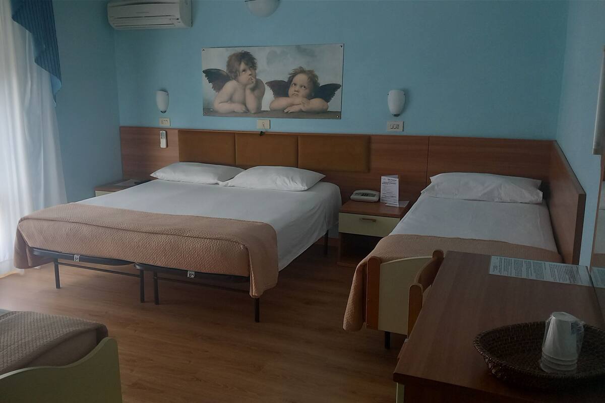 A hotel room with two beds, a wooden headboard, a painting of cherubs, a bedside table with a lamp, and a window with curtains.