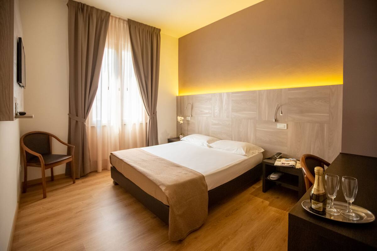 A hotel room with a bed, a wooden chair, a desk with a tray of champagne glasses, and a nightstand with a phone and a lamp.
