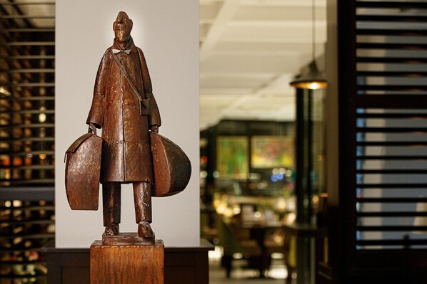 Una estatua de bronce de un hombre sosteniendo una maleta y un escudo, ubicada en un pedestal en un área de recepción con diseño interior moderno, que incluye un techo con iluminación empotrada y una partición de vidrio al fondo.
