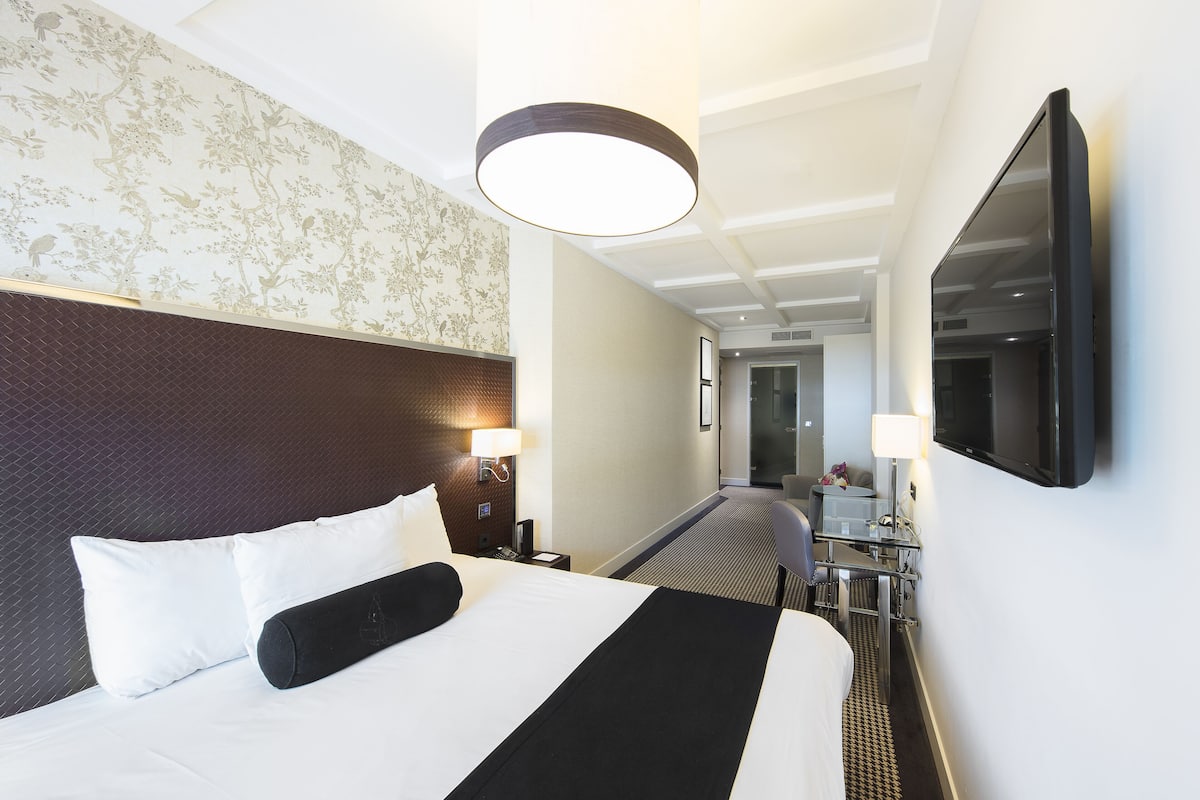A hotel room with a large bed, a desk, a chair, a TV, and a lamp.