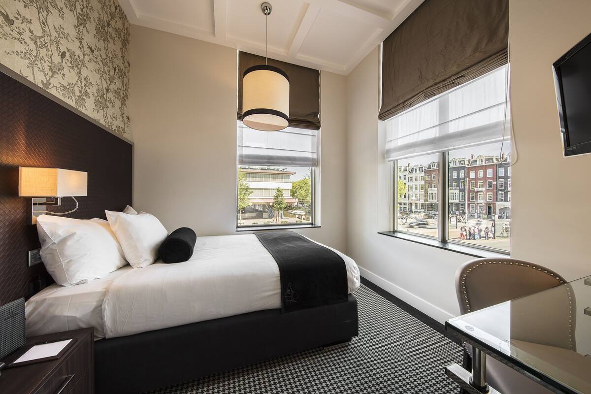 A hotel room with a bed, a desk, a chair, a lamp, and a window with a view of buildings.