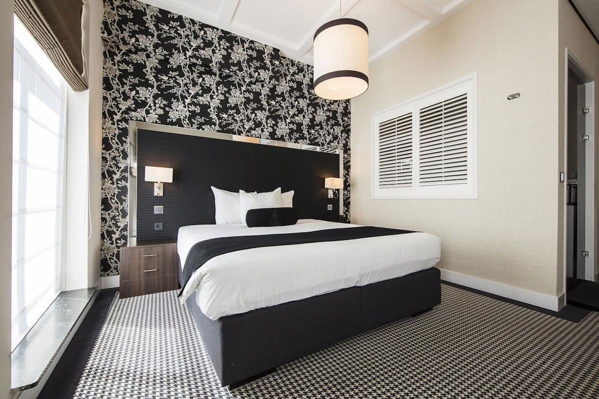 A hotel room with a large bed, a nightstand, a wall-mounted lamp, and a window with blinds.