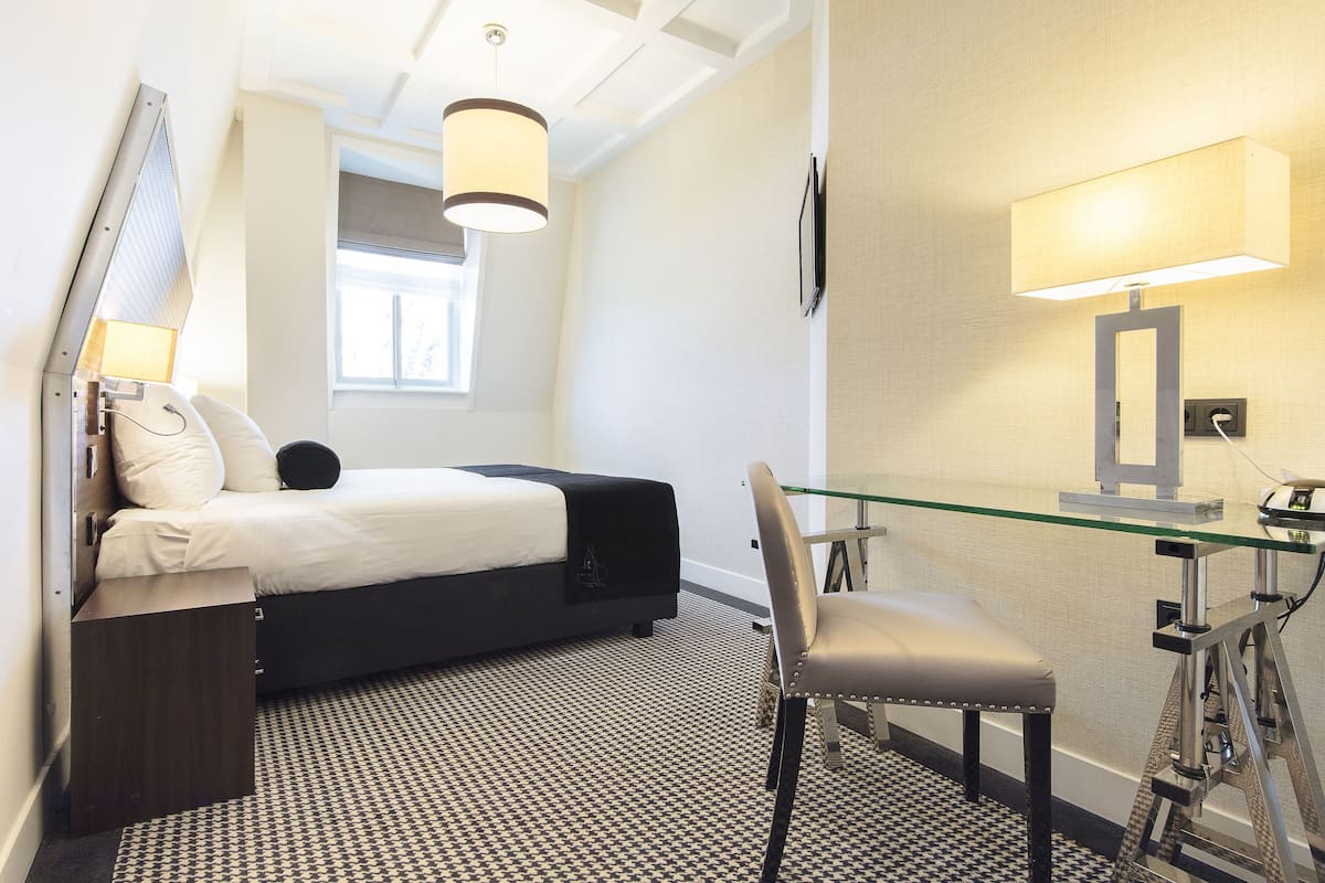 A hotel room with a bed, a desk with a chair, a lamp, and a window.