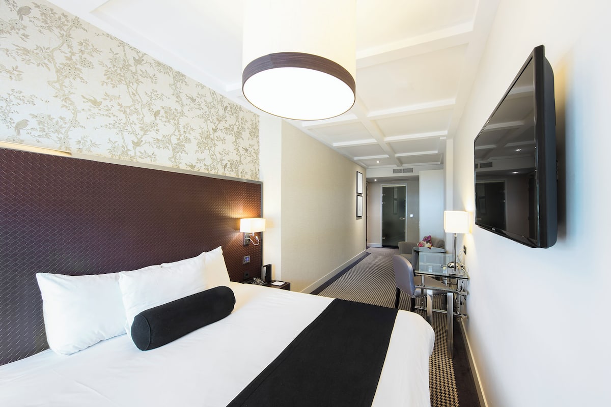 A hotel room with a large bed, a desk, a chair, a TV, and a lamp.