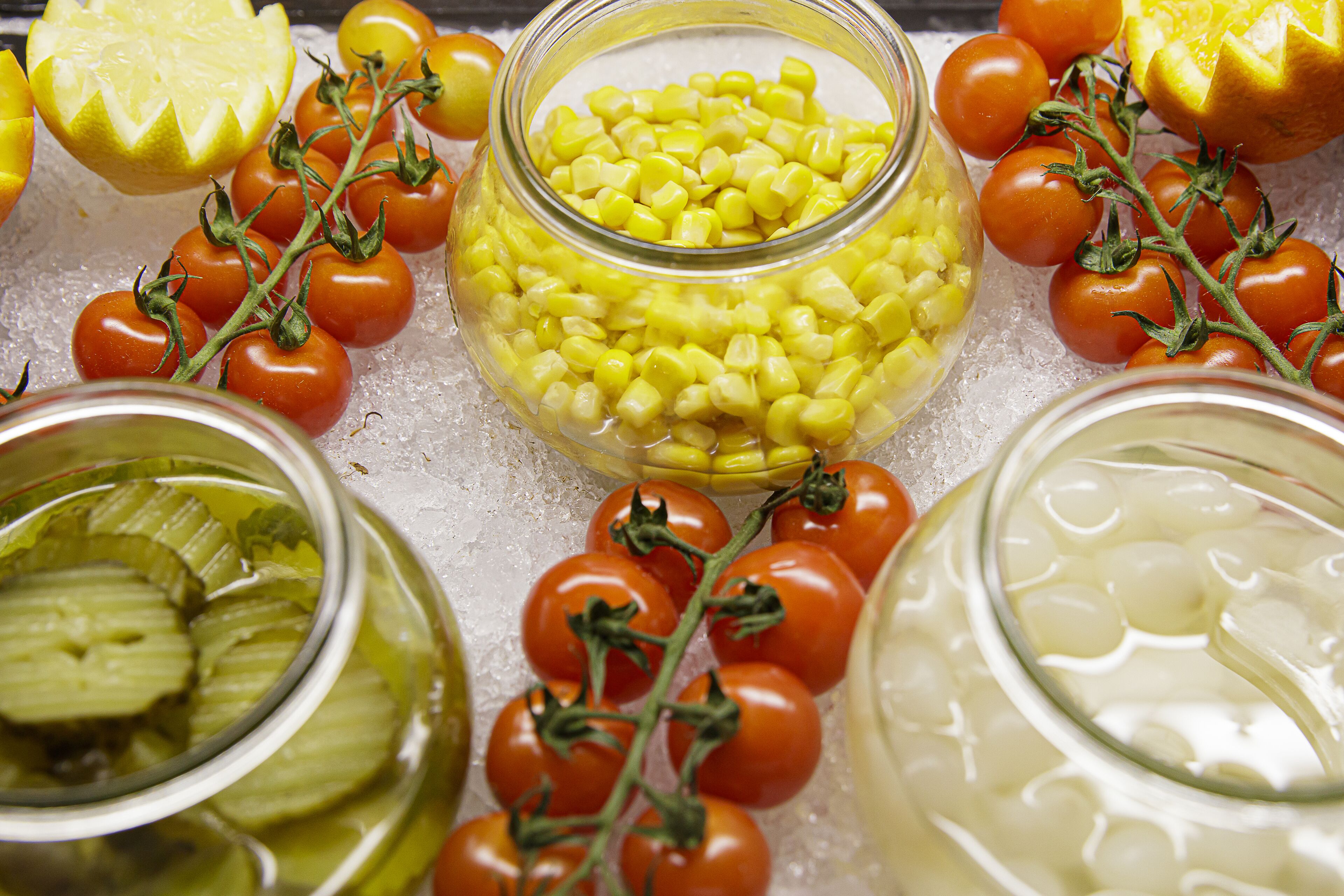 A variety of preserved vegetables including tomatoes, corn, and pickles on ice.