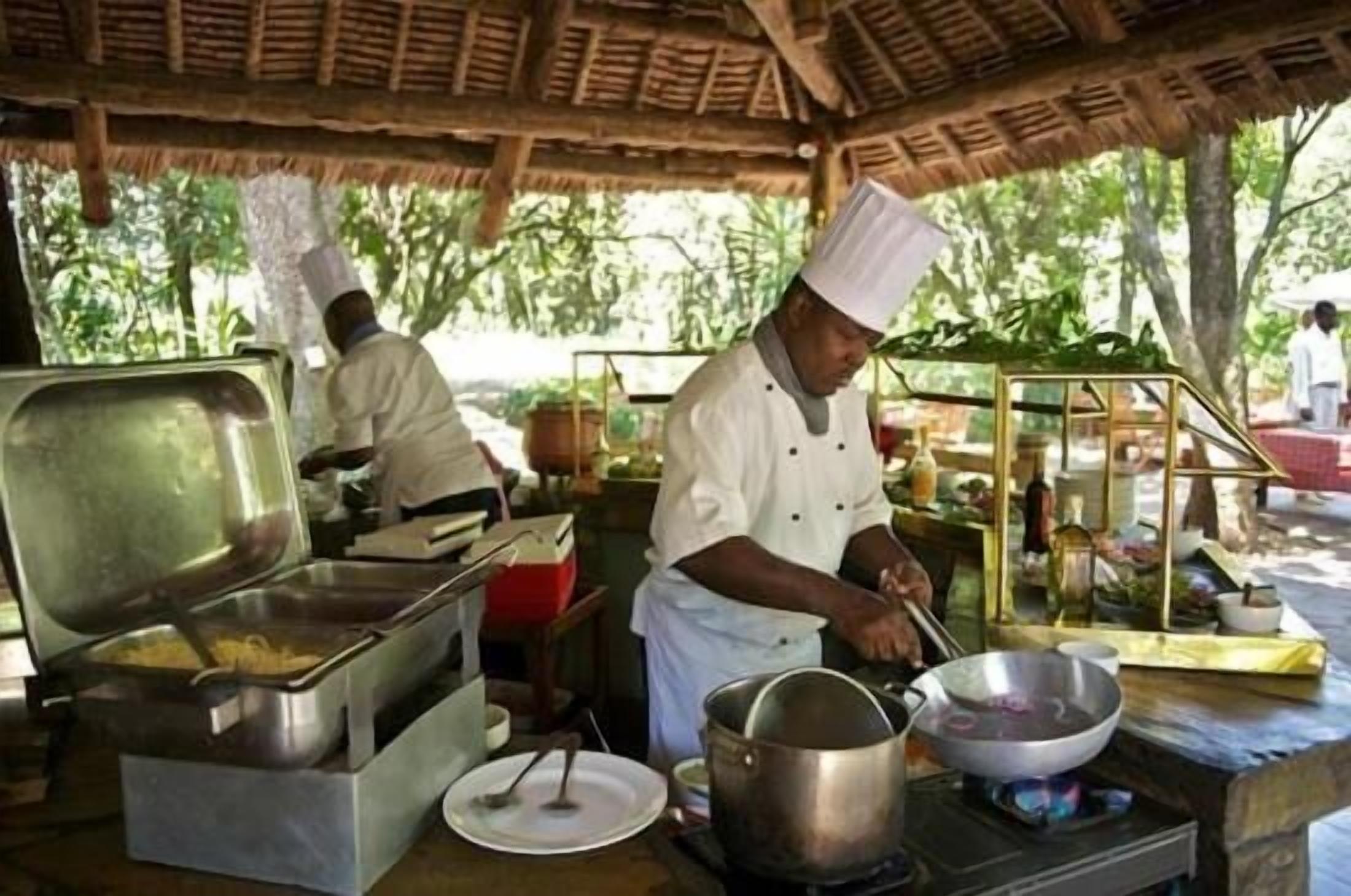 Uno chef prepara cibo in una cucina all'aperto con tetto in paglia, attrezzature da cottura in acciaio inossidabile e un'ampia scelta di ingredienti e utensili.