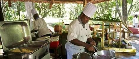 A chef preparing food in an outdoor kitchen with a thatched roof, stainless steel cooking equipment, and a variety of ingredients and utensils.
