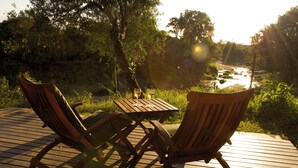 A wooden deck with two chairs and a small table, overlooking a river and surrounded by greenery. - Mara Intrepids Tented Camp (Maasai Mara)