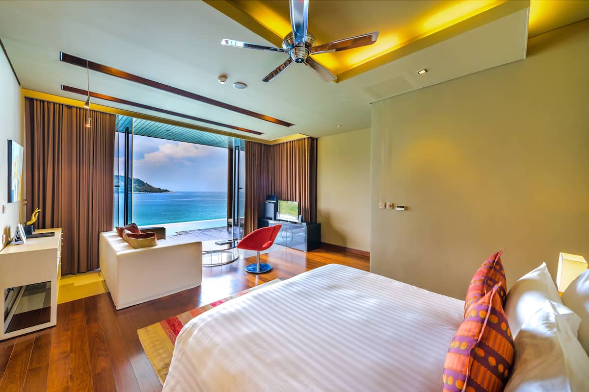 A modern hotel room with a large bed, a desk, a chair, and a TV. The room has a view of the sea.
