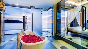 A modern bathroom with a large bathtub filled with rose petals, a glass shower enclosure, and a view of the sea. - Impiana Private Villas Kata Noi (Karon)