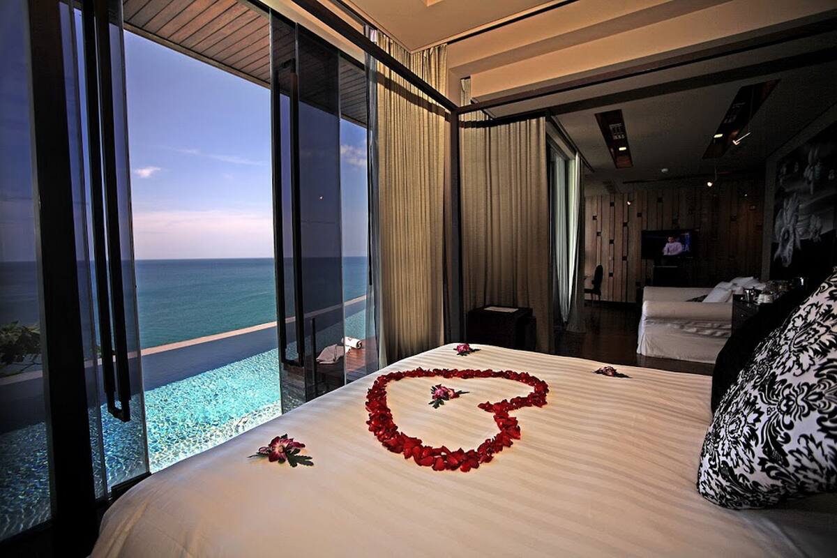 A bedroom with a large bed, a heart-shaped arrangement of red flowers on the bed, and a view of the ocean through floor-to-ceiling windows.