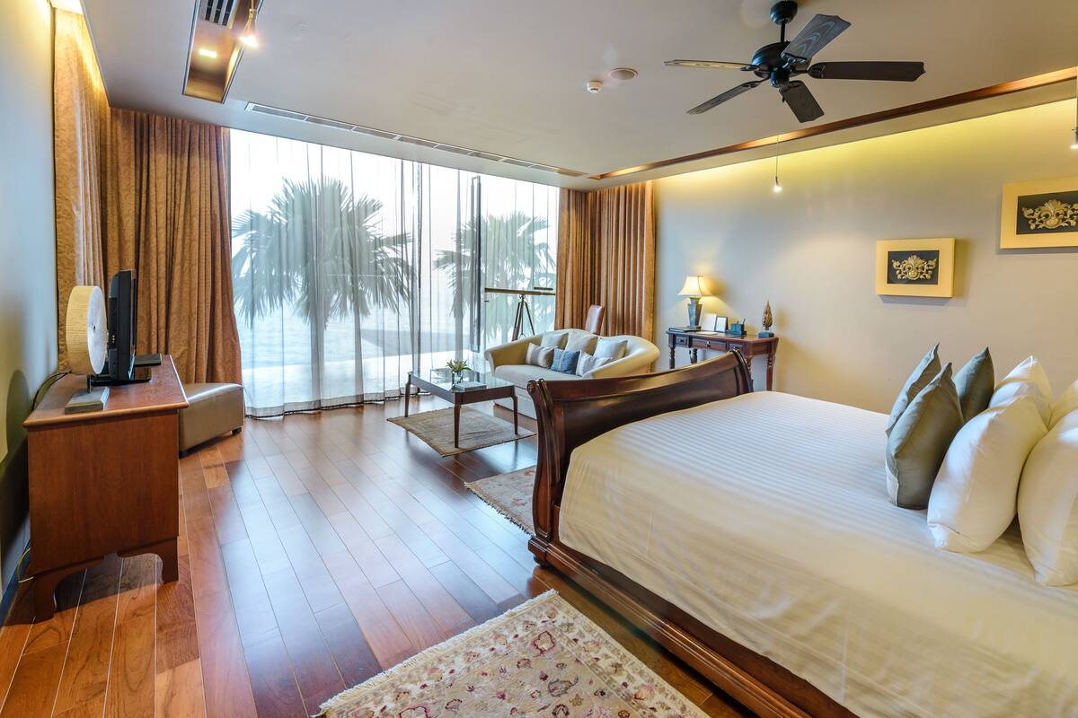 A hotel room with a large bed, a desk, a chair, a ceiling fan, and a view of palm trees through the window.
