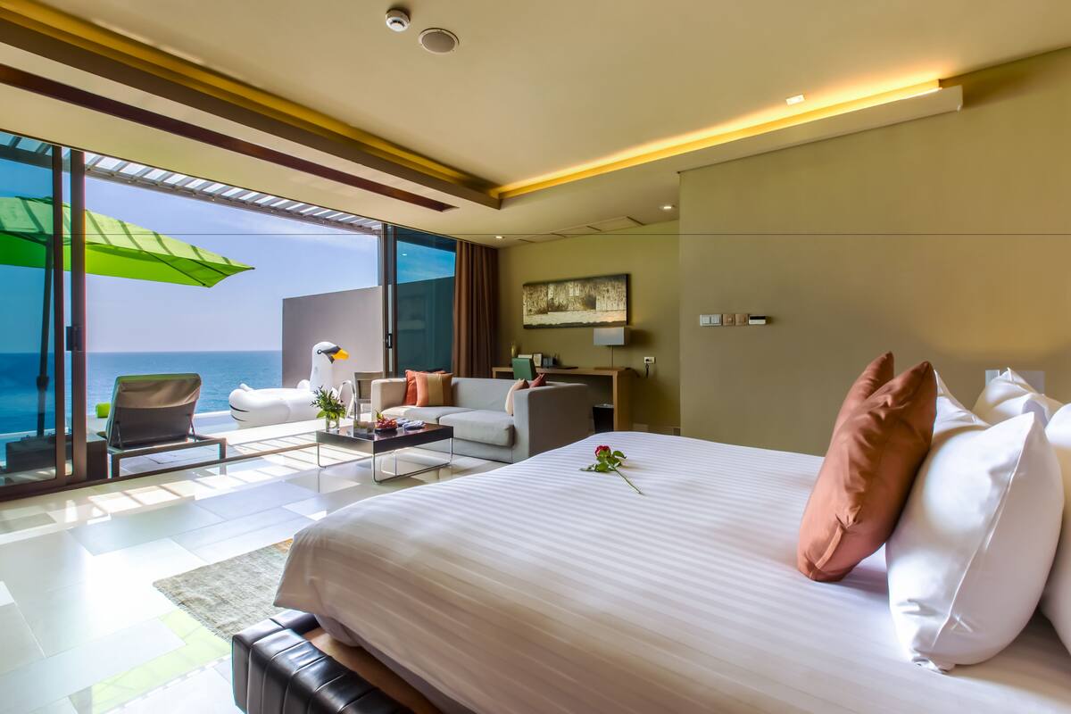 A modern hotel room with a large bed, a sofa, a desk, and a view of the ocean.