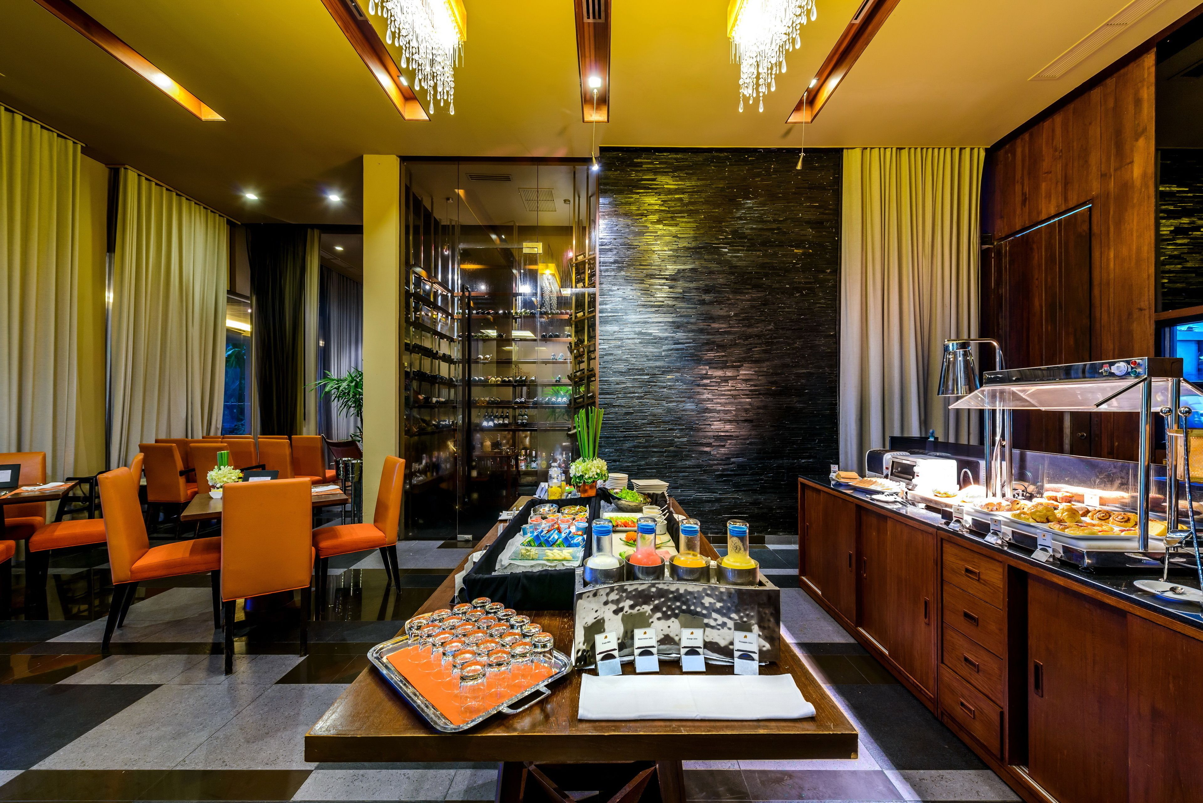 A hotel dining area with a buffet, orange chairs, and a long table with various food items.