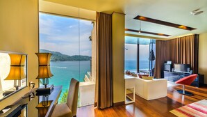 A modern hotel room with a large window offering a view of the sea and hills. It includes a desk with lamps, a sofa, a TV, and a red chair. - Impiana Private Villas Kata Noi (Karon)