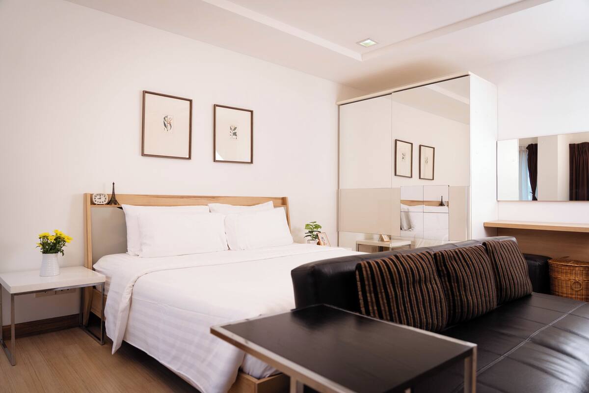 A modern hotel room with a bed, a sofa, a small table, and a nightstand. There are framed pictures on the wall and a mirror above the sofa.