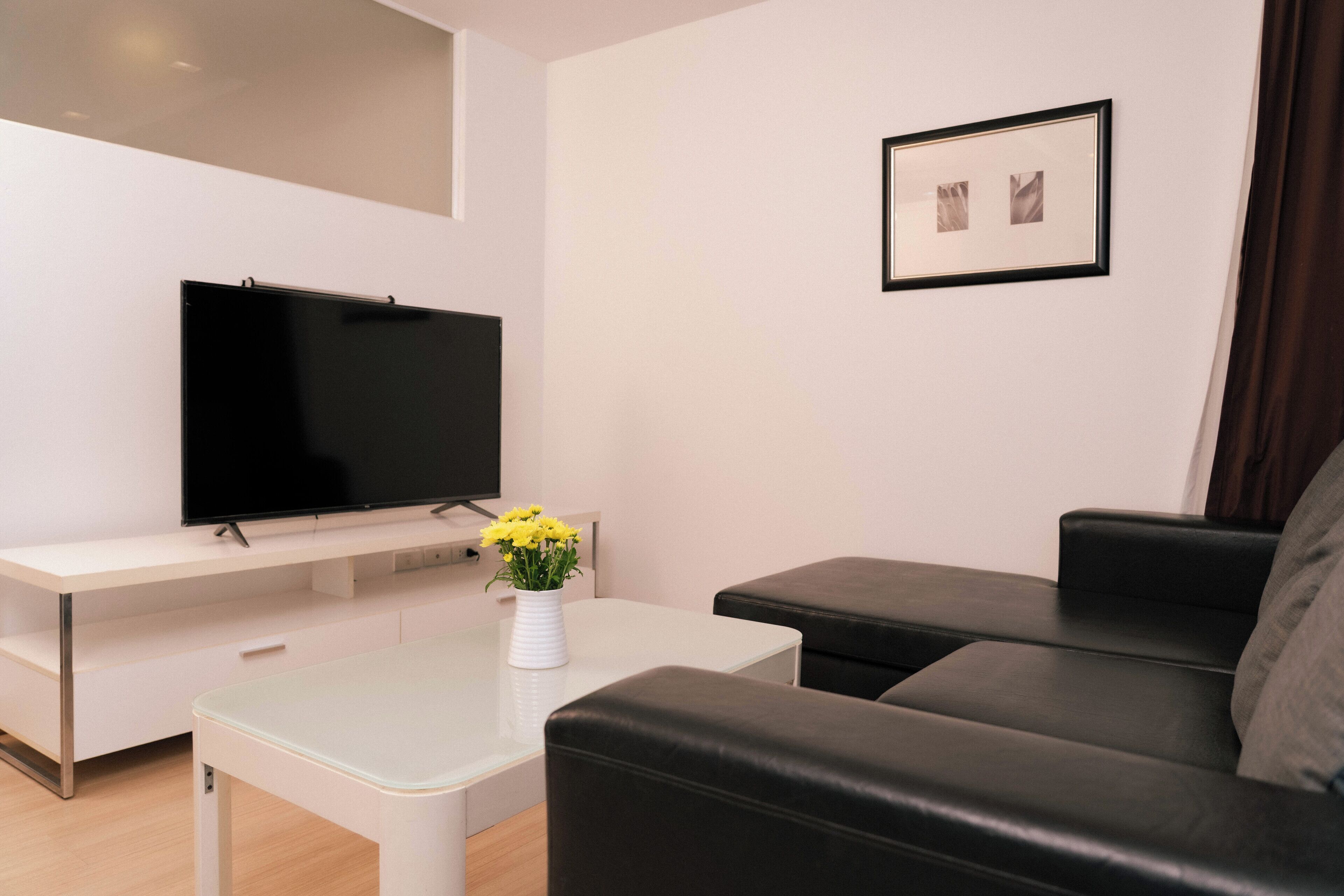 A modern living room with a flat-screen TV, a white coffee table with a vase of yellow flowers, a black leather sofa, and a framed picture on the wall.