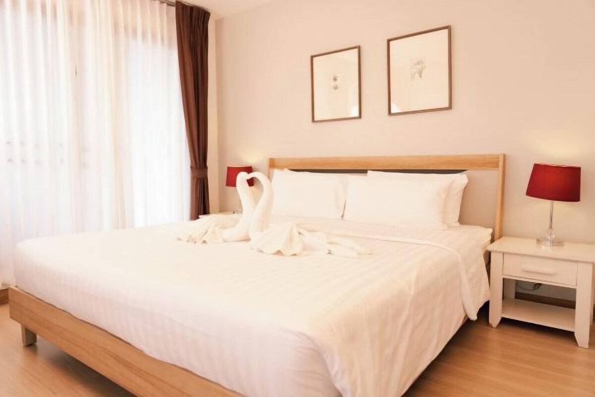 Un lit bien fait, avec du linge de lit blanc et un arrangement d’oreillers en forme de cygne. On y trouve une table de chevet avec une lampe rouge et un tableau encadré accroché au mur.
