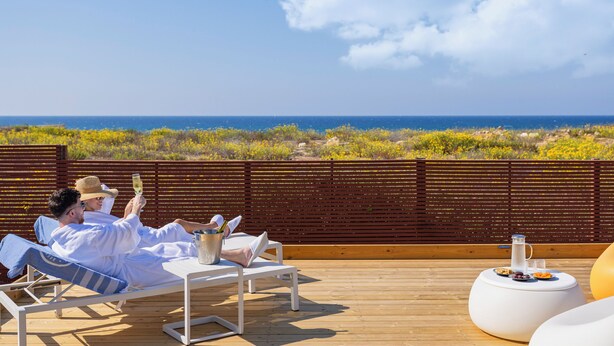 A couple relaxing on a wooden deck with a view of the ocean and yellow flowers.