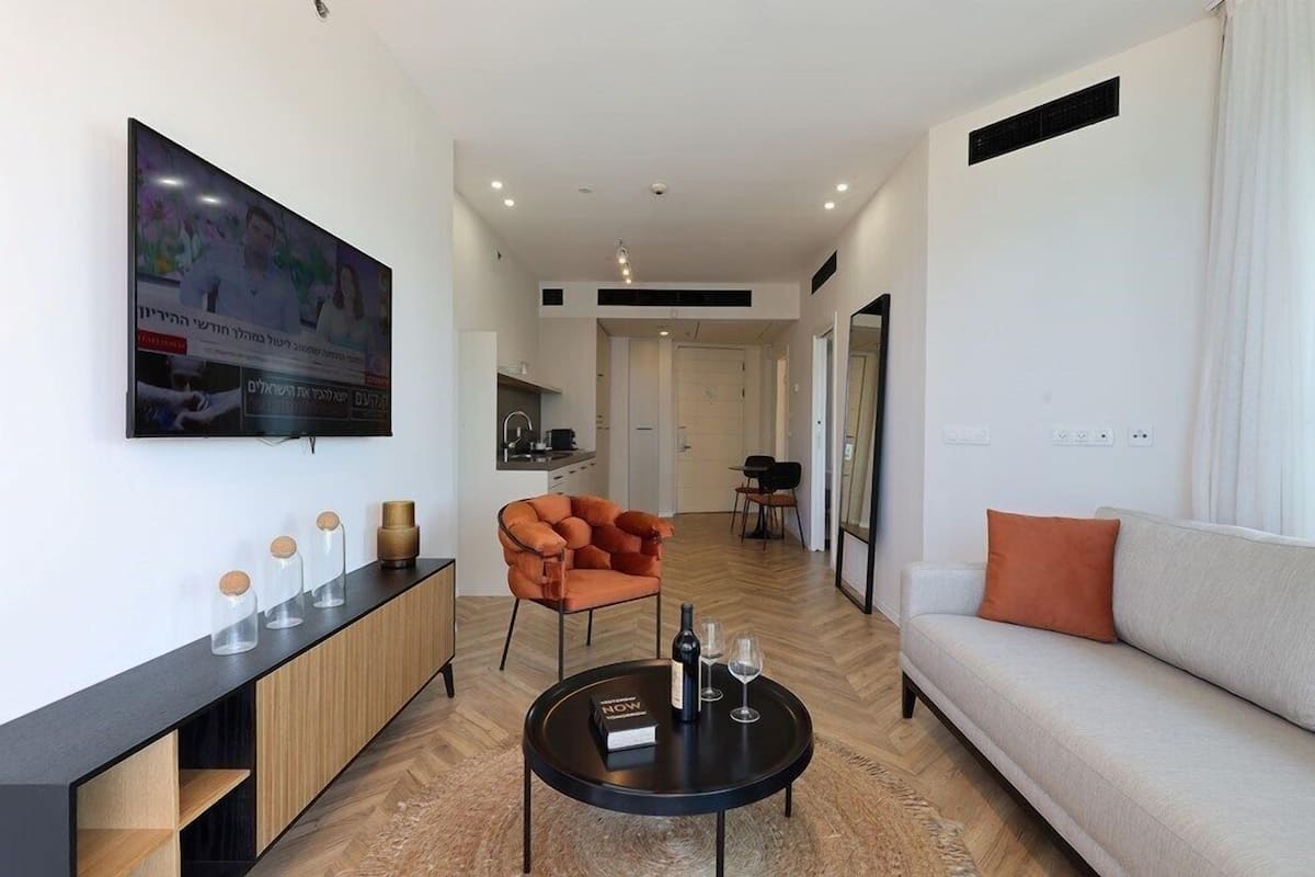 A modern living room with a sofa, armchair, and coffee table. There is a TV mounted on the wall and a cabinet with jars.