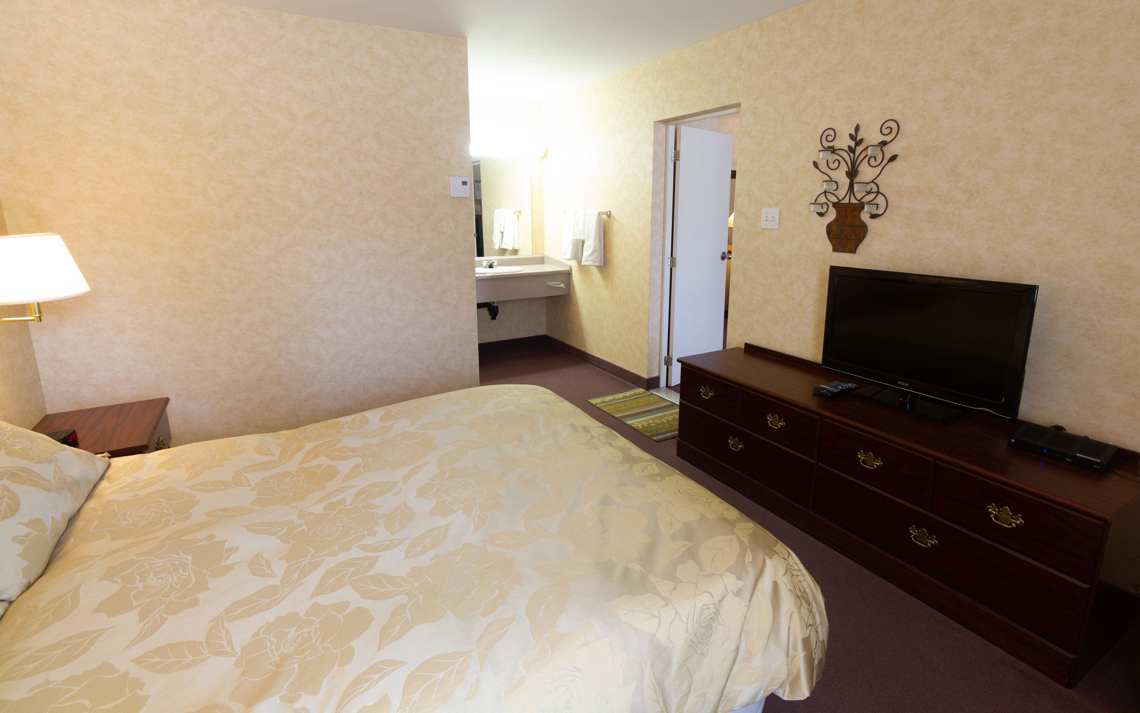 A hotel room with a large bed, a dresser with a flat-screen TV, a vanity with a mirror, and a wall-mounted light fixture.
