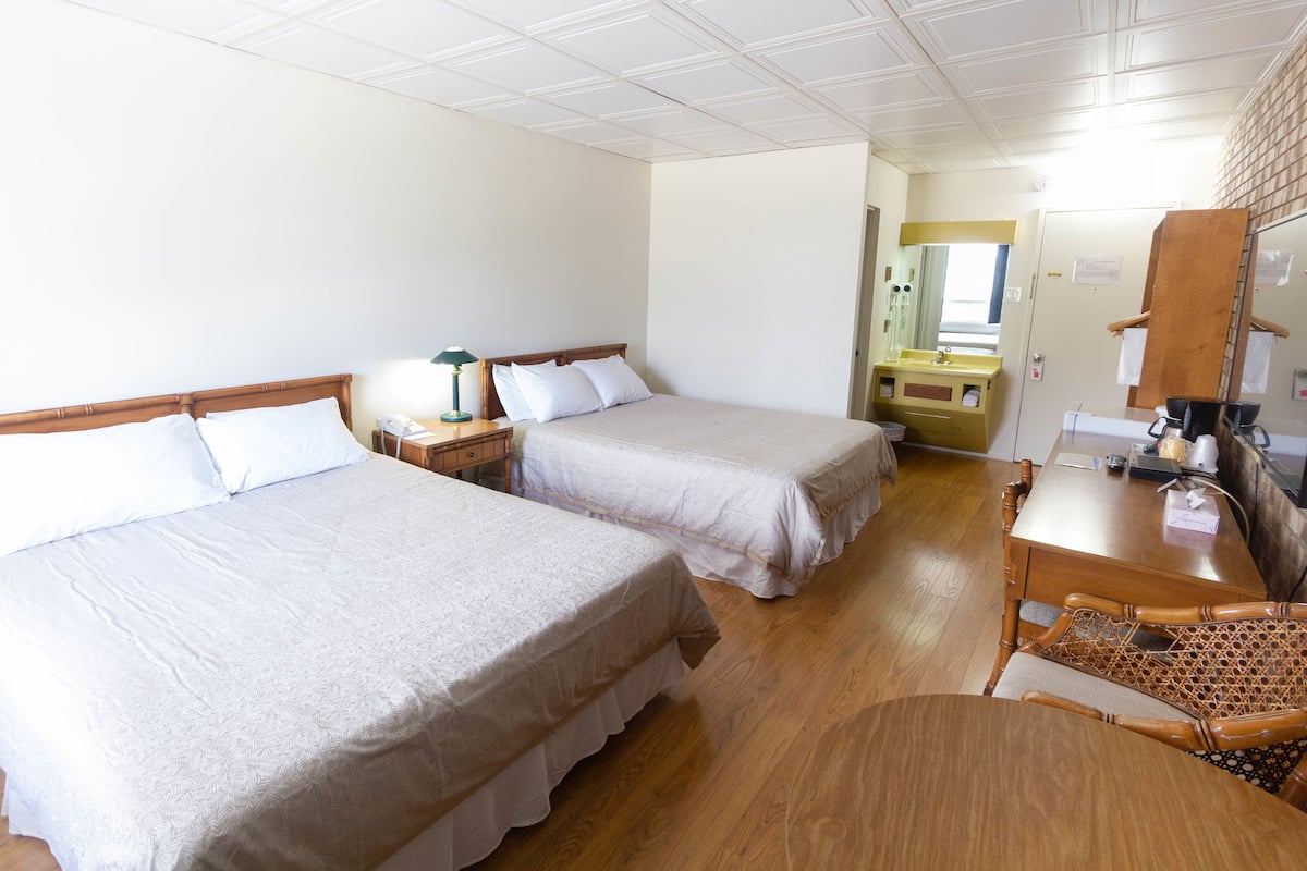 Habitación de hotel con dos camas, un escritorio de madera, un espejo, un lavamanos y una pequeña mesa con una cesta.
