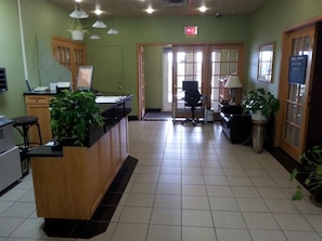 A reception area with a wooden desk, a computer, a telephone, and a plant. There is a door leading to another room.
