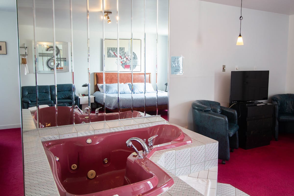 Habitación de hotel moderna con una bañera roja grande, una cama con cabecero de madera, un televisor de pantalla plana y una zona de estar con dos sillones.