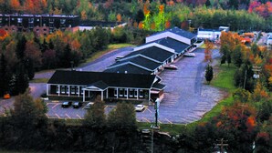 A multi-story hotel with a parking lot and a view of a forested area with autumn foliage. - Auberge La Rocaille (Shawinigan)