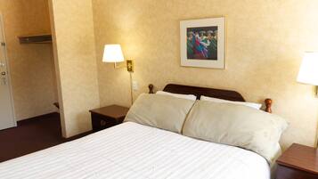 Une chambre d’hôtel avec un grand lit, deux tables de chevet, une petite table, une lampe fixée au mur et un tableau encadré accroché au mur.