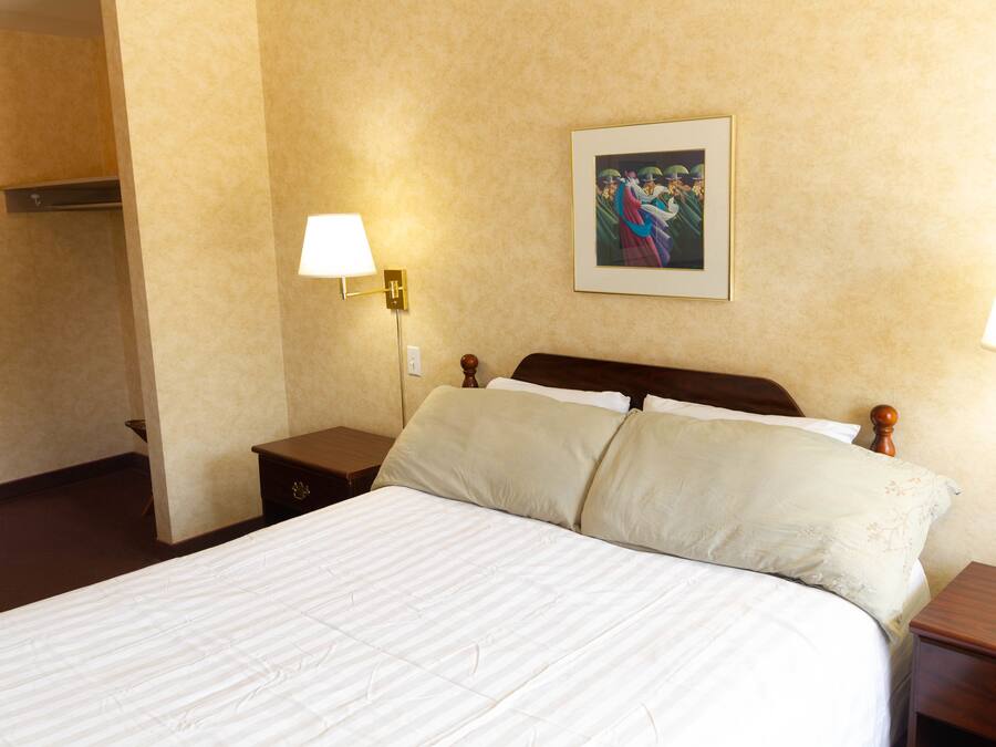 Une chambre d’hôtel avec un grand lit, deux tables de chevet, une petite table, une lampe fixée au mur et un tableau encadré accroché au mur.