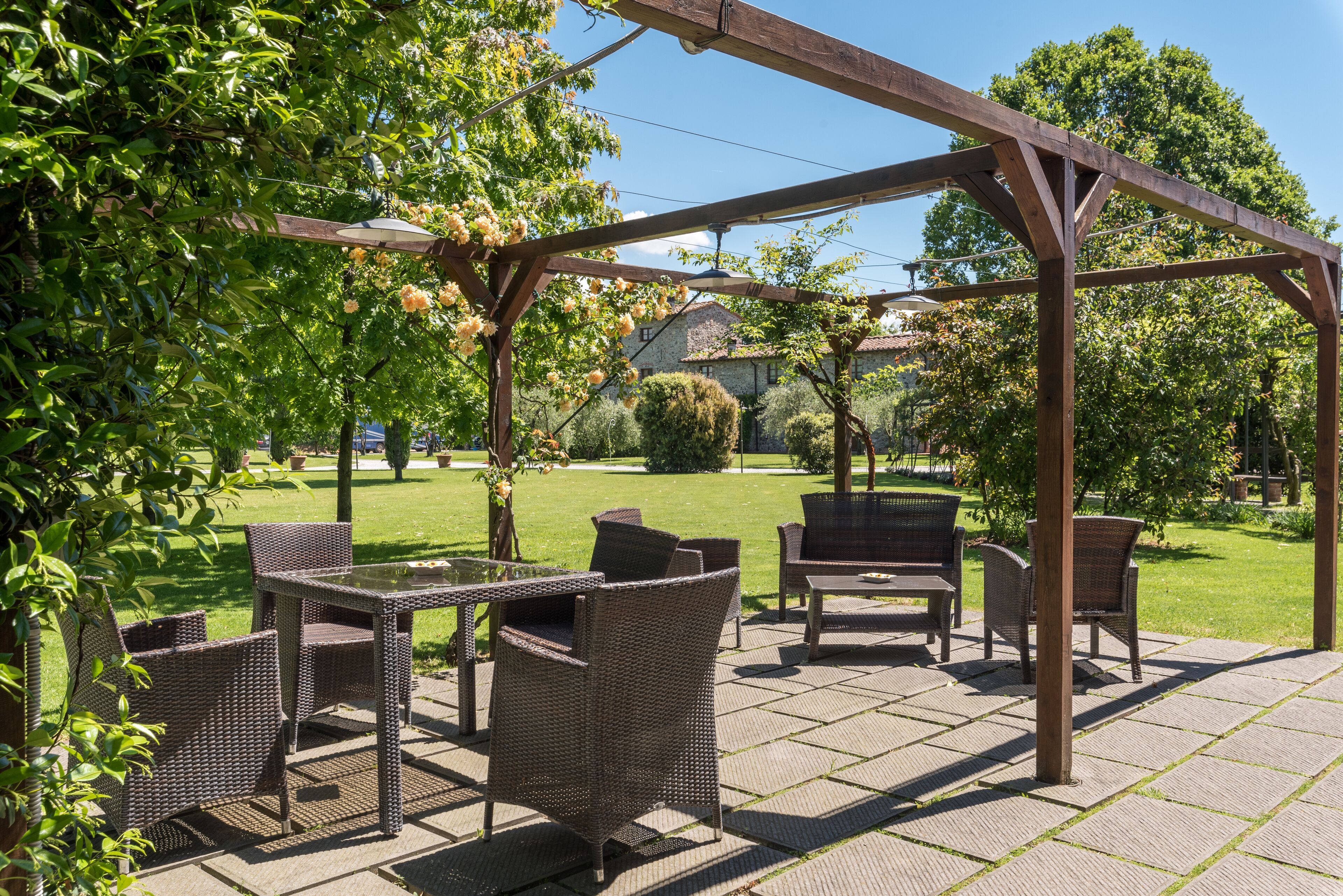 Un patio exterior con mobiliario de mimbre, una pérgola y vistas a un jardín y una casa al fondo.
