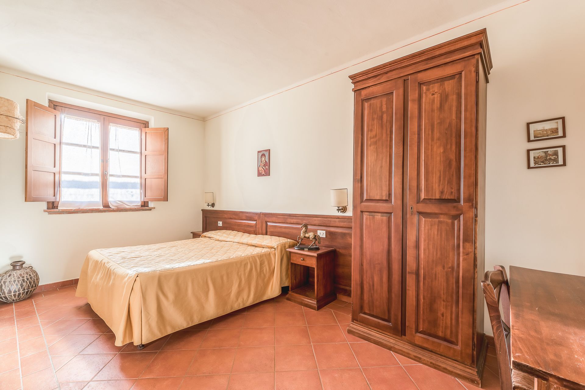 A bedroom with a bed, a nightstand, a wooden wardrobe, a window with curtains, and a wall-mounted lamp.