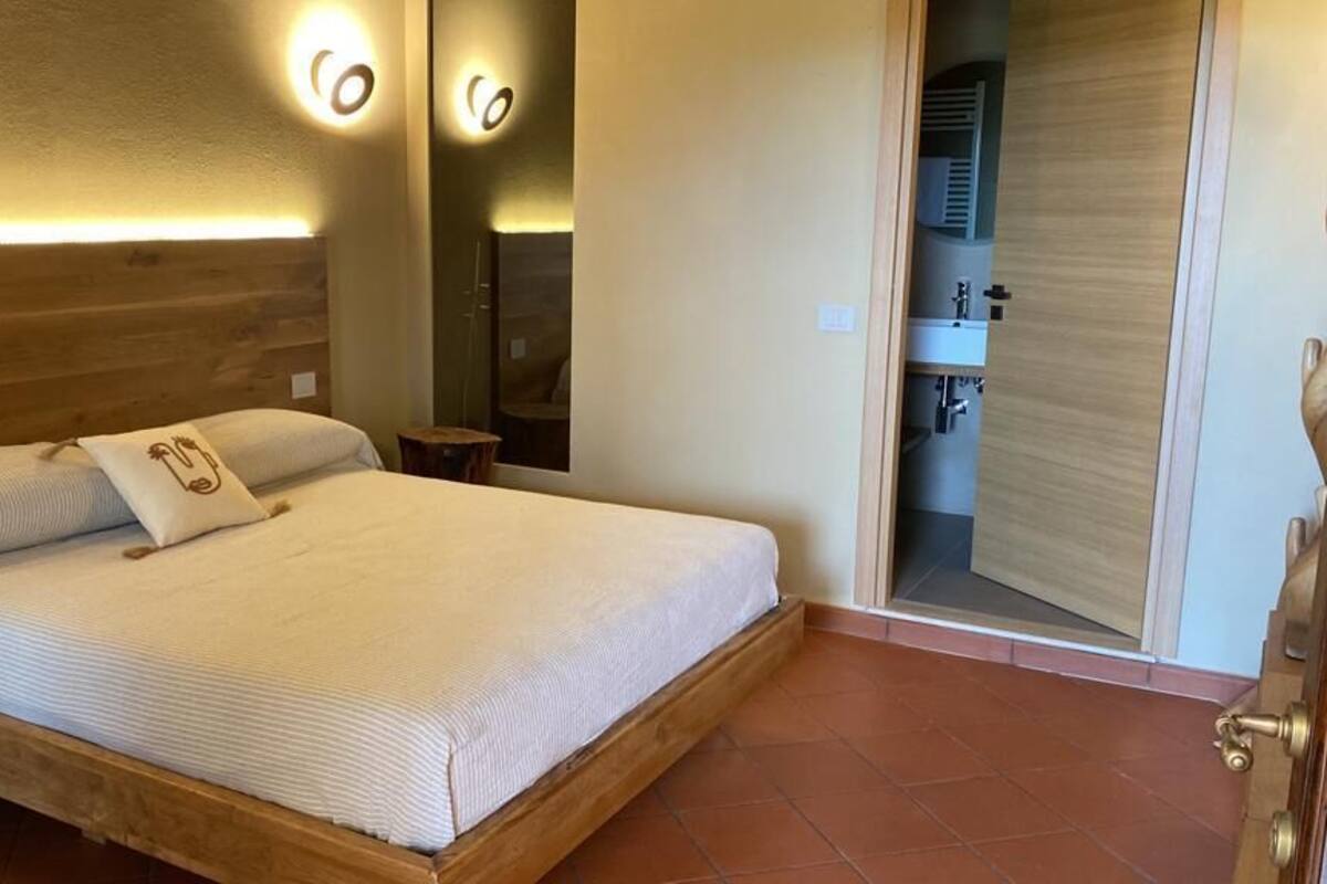 Un dormitorio con una cama de madera, una mesita de noche, un espejo y un baño visible a través de una puerta abierta.