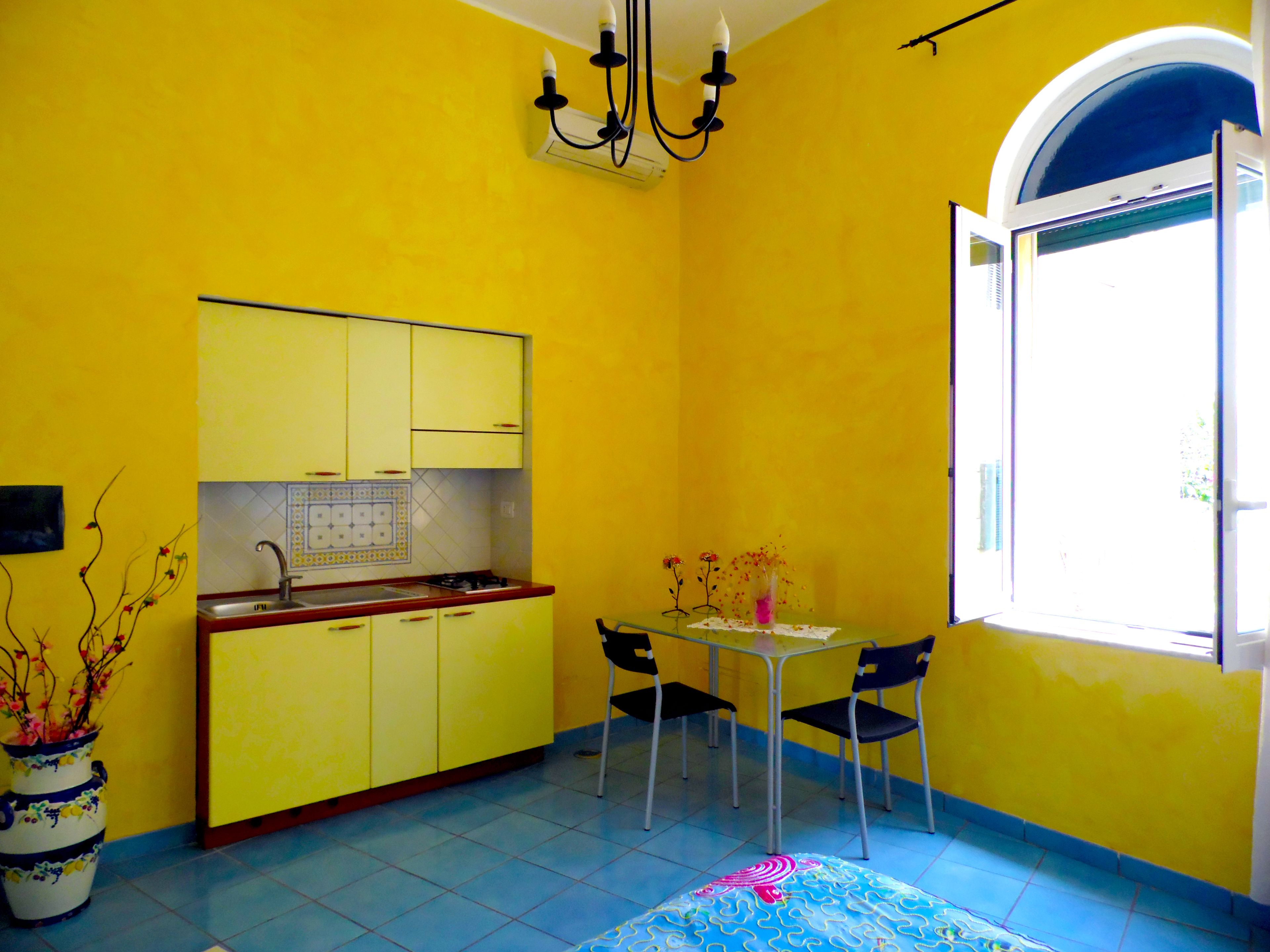 A small kitchen with yellow cabinets, a sink, and a stove. There is a dining table with chairs and a vase with flowers.