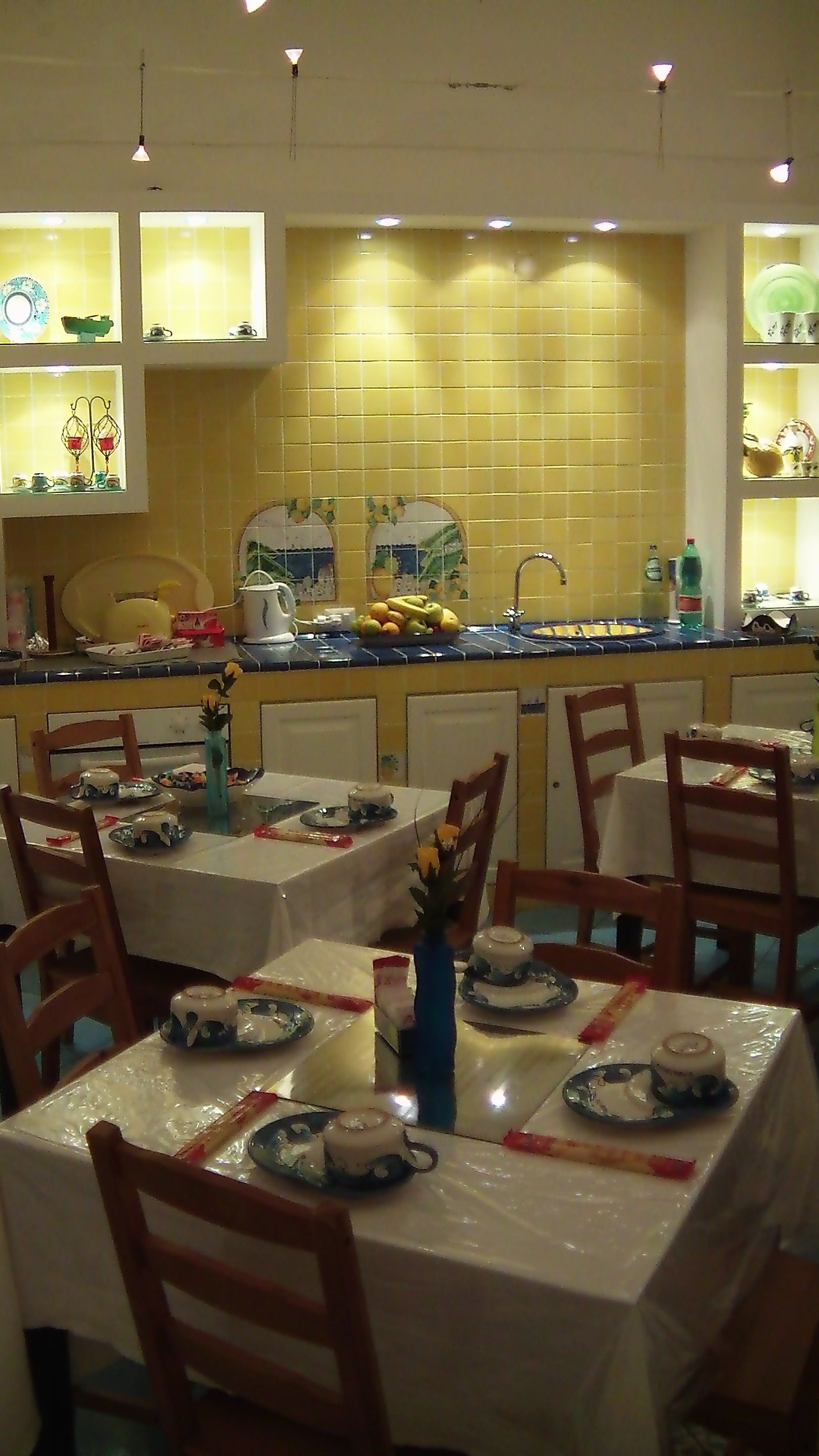 A restaurant with tables set for dining, a kitchen area, and shelves with kitchenware.