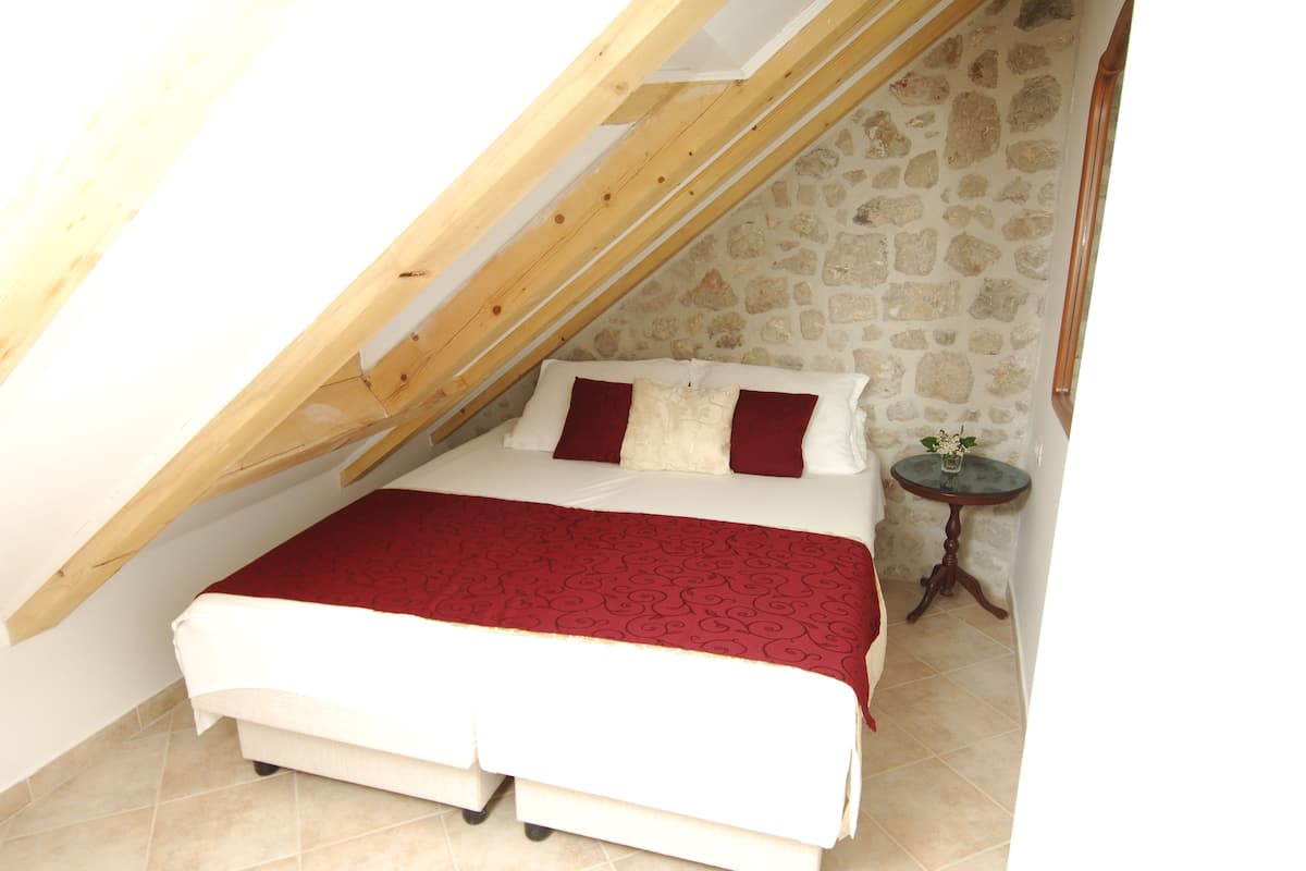 Une chambre avec un plafond en pente, un lit recouvert d’une courtepointe rouge et blanche, une petite table ronde ornée d’un vase de fleurs et un mur en pierre.