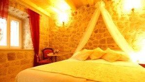 Una habitación con pared de piedra, una cama con ropa de cama blanca, una silla roja y una mesita con un florero.