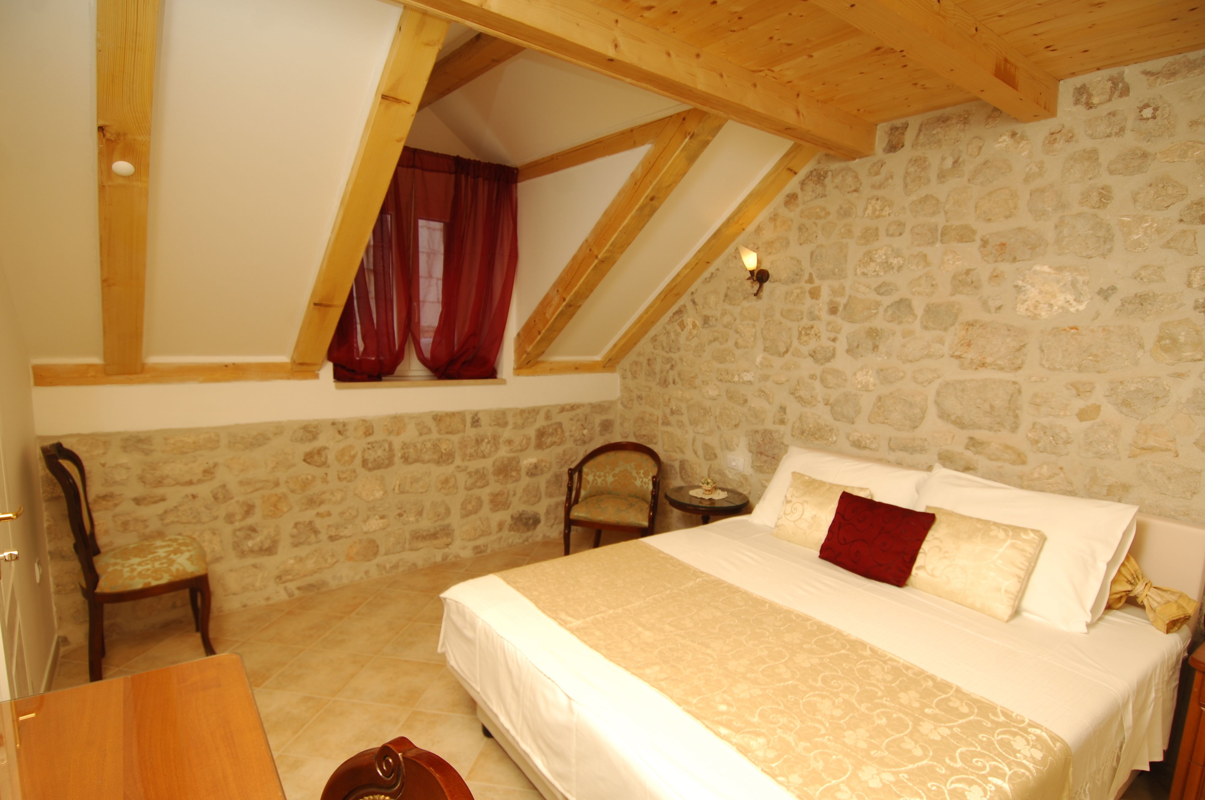 A bedroom with a stone wall, a wooden ceiling, a bed with white and red bedding, and a chair.