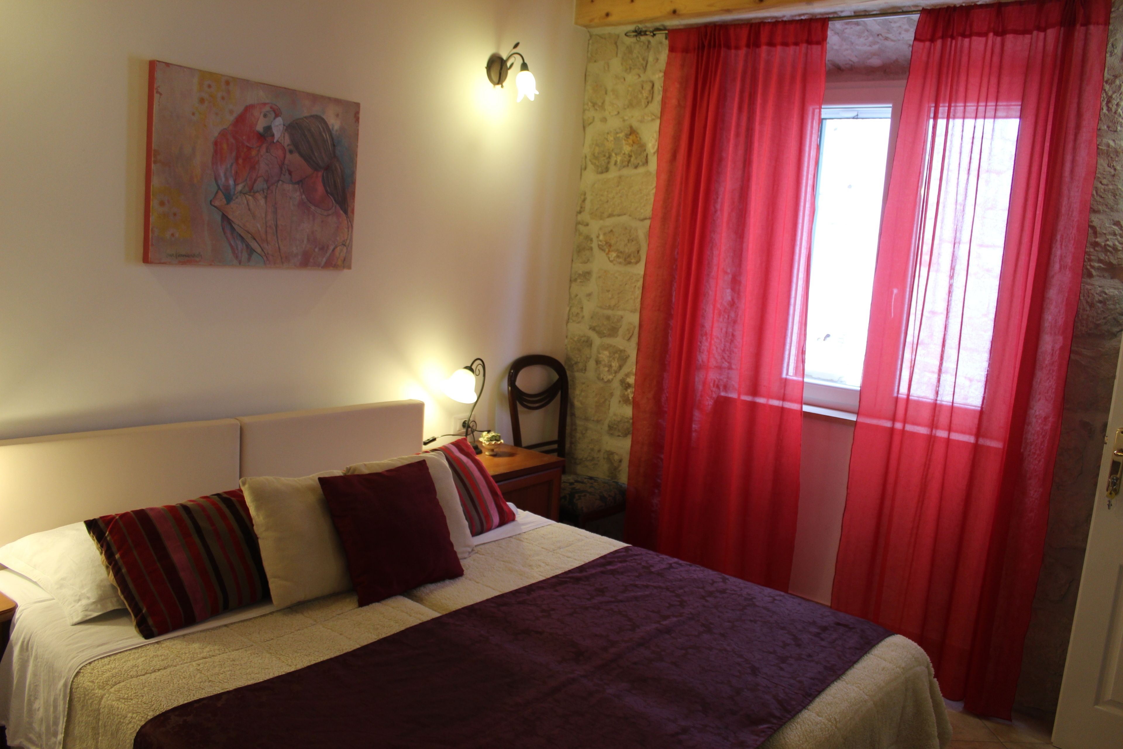 A bedroom with a bed, a nightstand, a lamp, a painting on the wall, and a window with red curtains.
