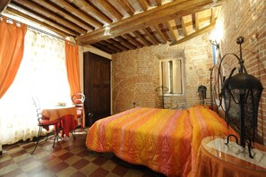 Ein Zimmer mit einem Bett, einem Tisch und einem Stuhl. Die Wände sind aus Stein und die Decke aus Holz.