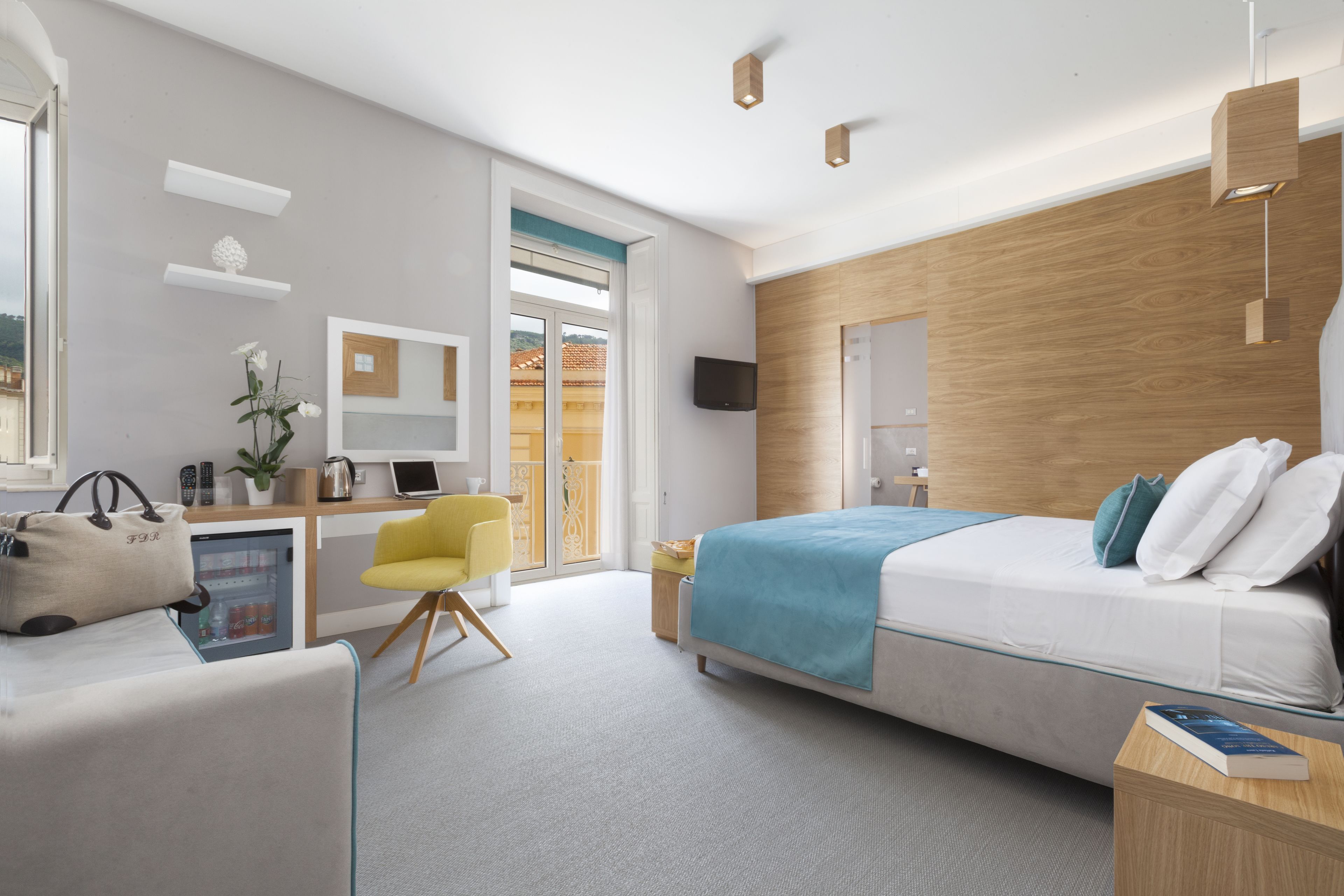 A modern hotel room with a bed, a desk with a computer, a chair, and a small refrigerator.