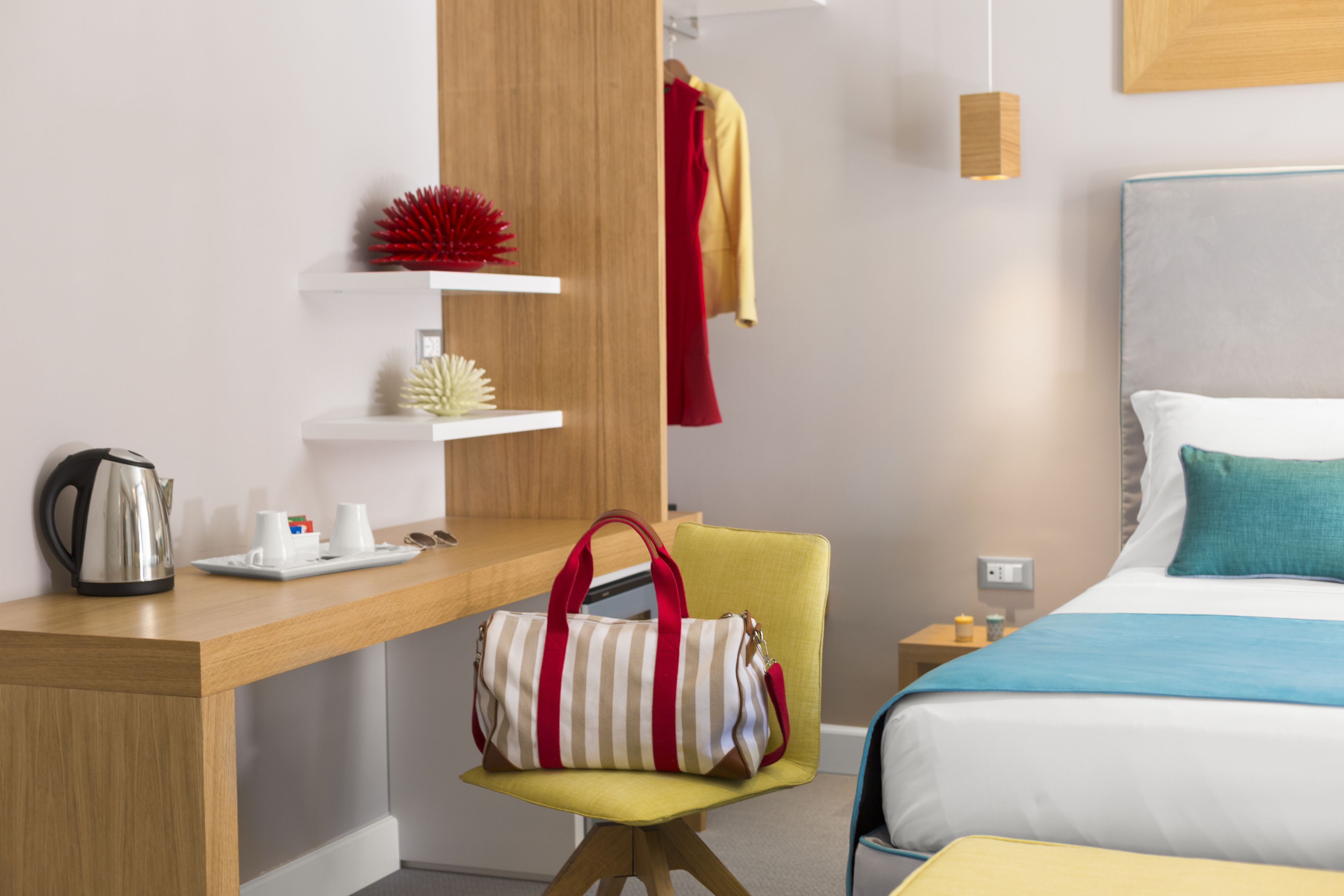 A hotel room with a bed, a desk with a kettle, a chair, and a bedside table with a lamp.