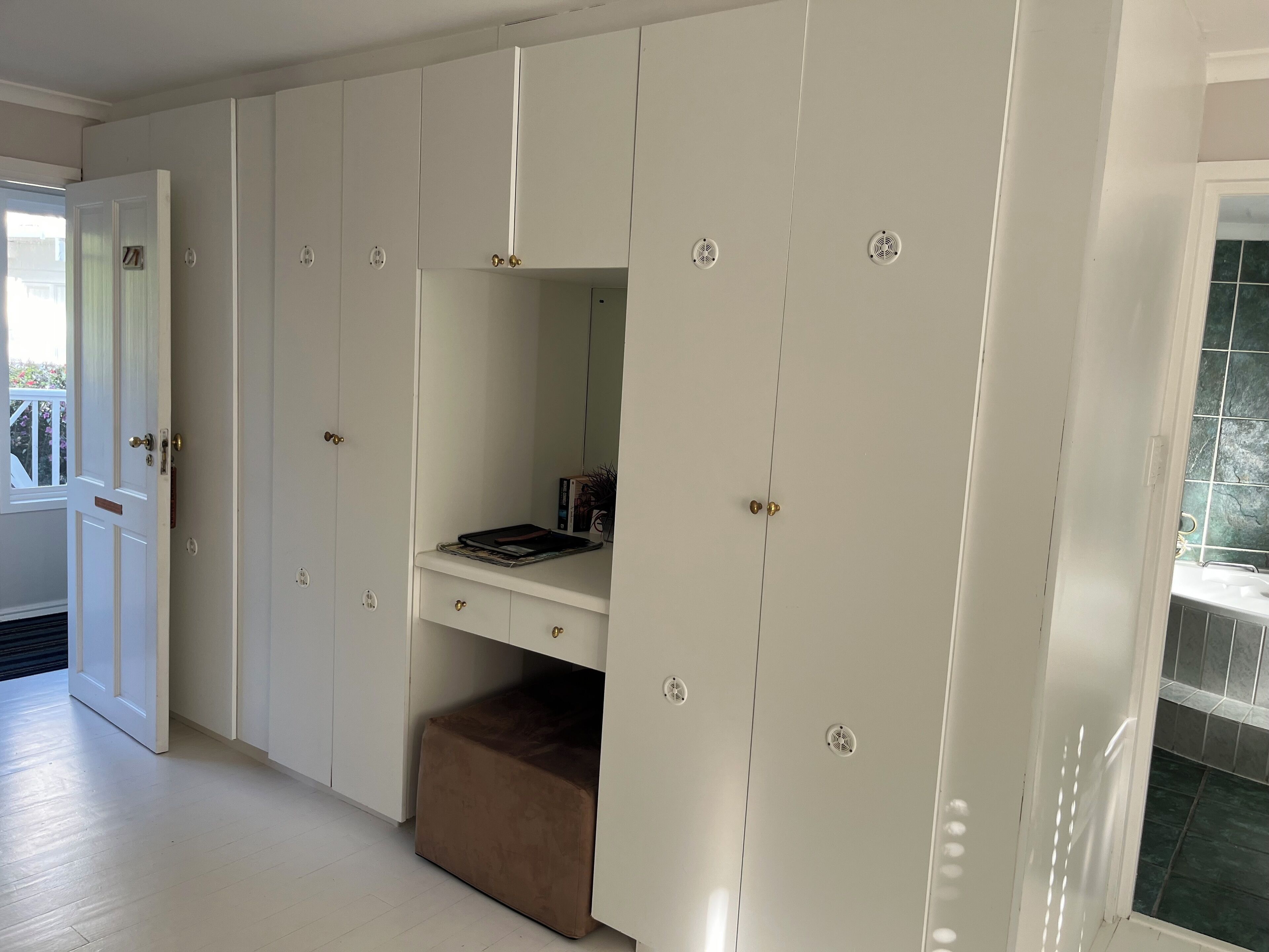 A room with a white wardrobe, a desk with a mirror, and a brown ottoman.
