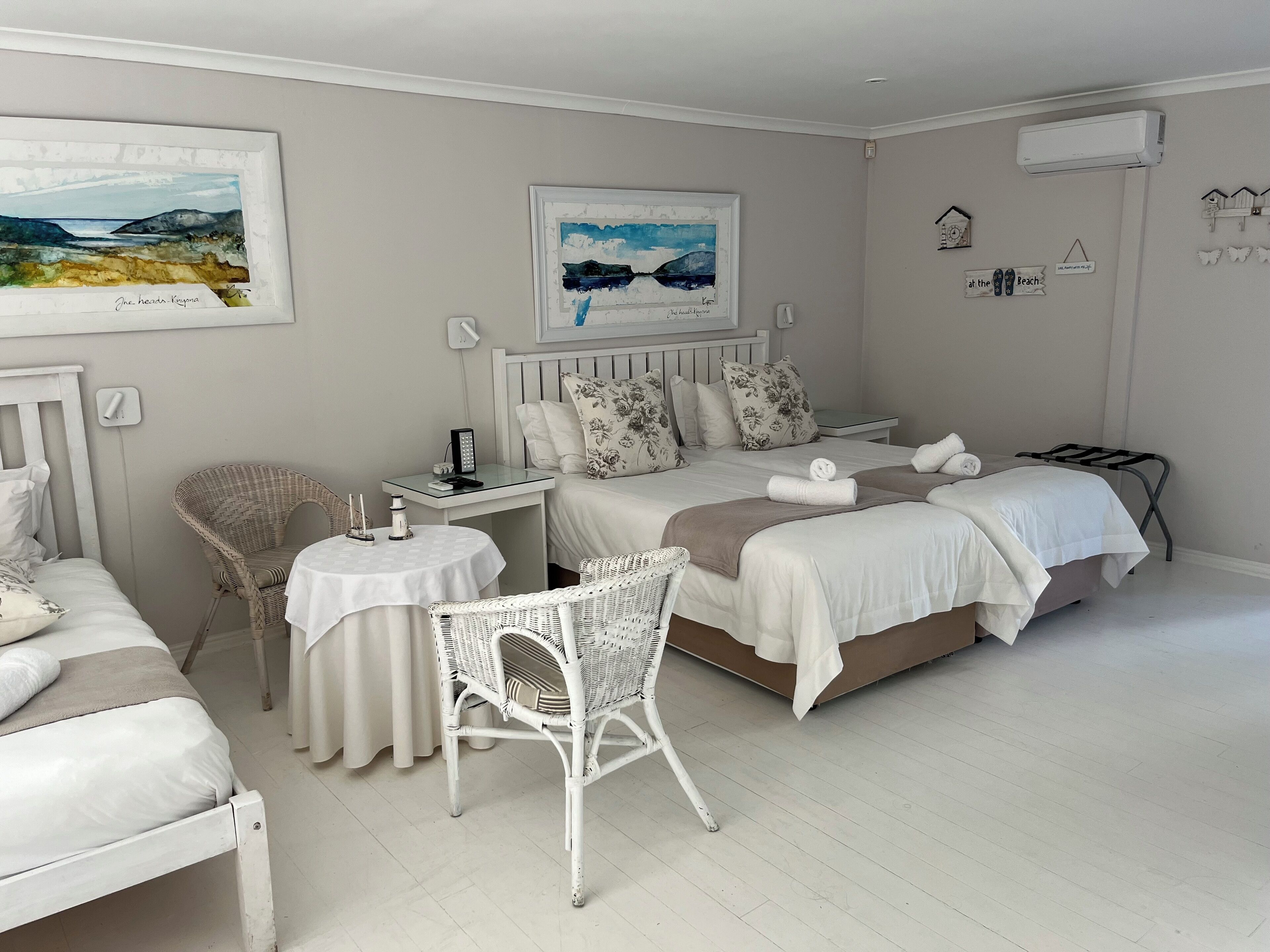 A hotel room with two beds, a small table, wicker chairs, and artwork on the walls.