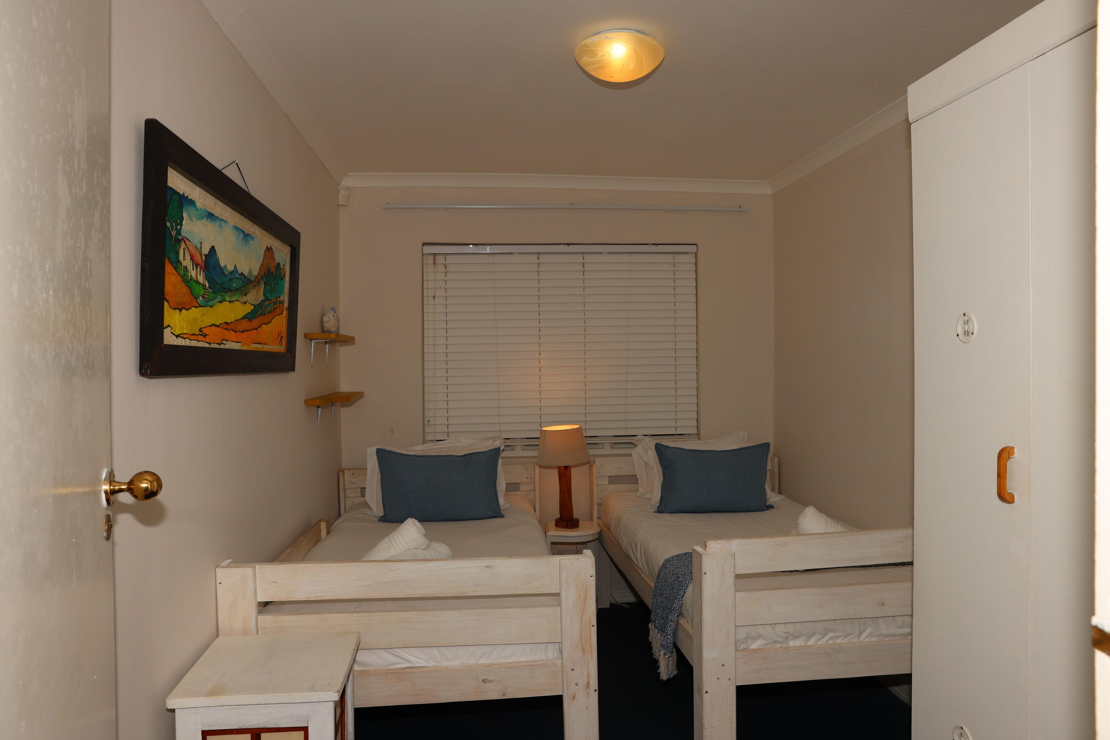 A room with two wooden beds, a window with blinds, a painting on the wall, and a lamp on a bedside table.
