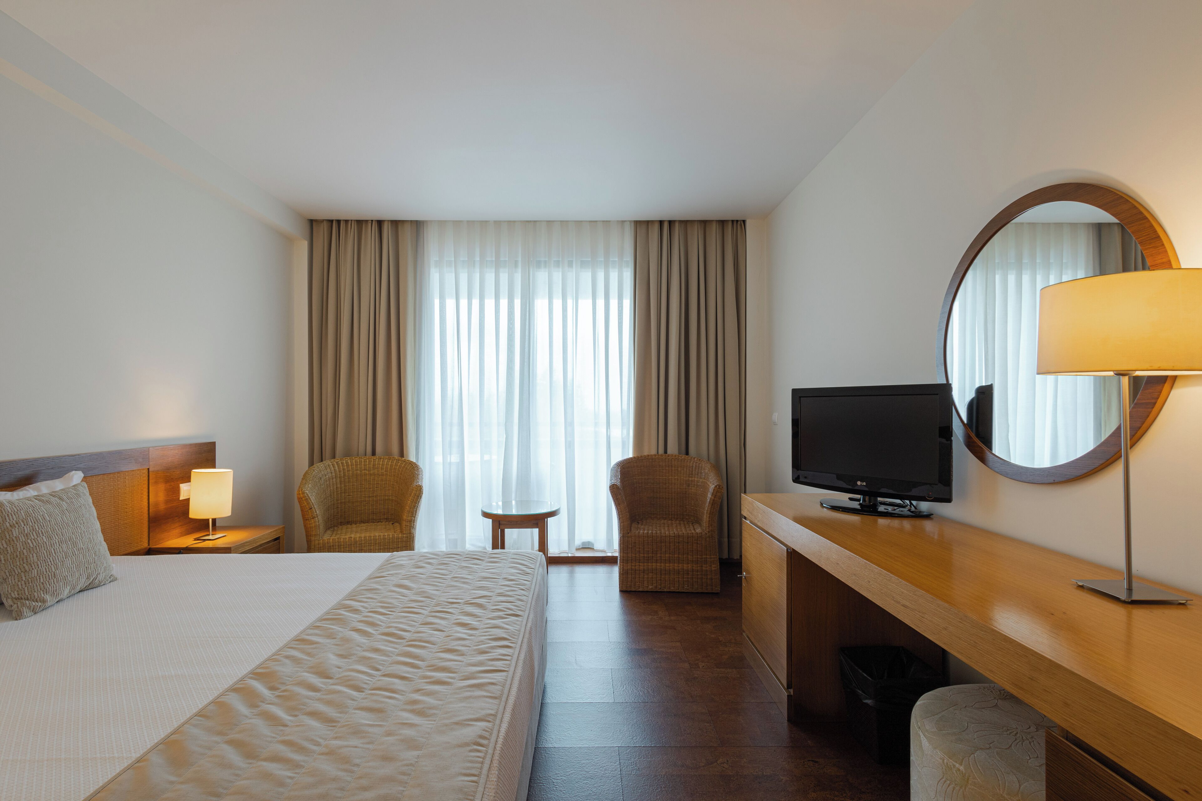 Ein Hotelzimmer mit einem Bett, zwei Sessel, einem Schreibtisch mit Fernseher, einer Lampe, einem kleinen Tisch und einem großen Fenster mit Vorhängen.